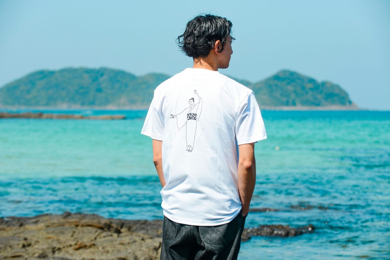 海を背に、踊り子のバックプリントTシャツを着た男性